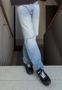 Personne portant un jean déchiré bleu clair et des baskets Nike noires, debout avec les jambes croisées sur un palier d'escalier à l'intérieur.