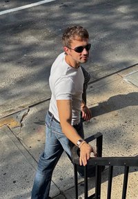 Un bărbat îmbrăcat cu un tricou alb ajustat și blugi albaștri, sprijinit de o balustradă neagră în aer liber, purtând ochelari de soare și un ceas de mână.