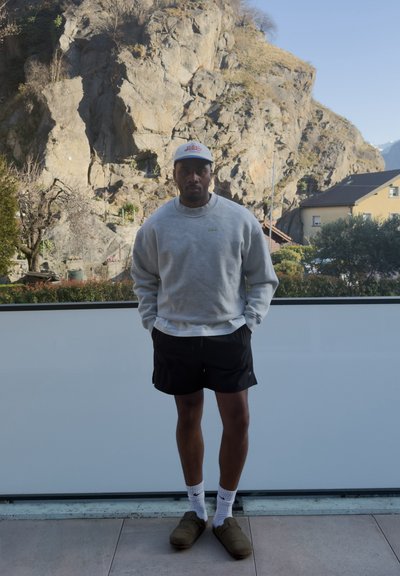 Hombre con sudadera gris, pantalones cortos negros, calcetines blancos, zapatos marrones y gorra blanca, de pie en una terraza con una colina rocosa y casas al fondo.