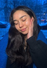 Jeune femme aux longs cheveux foncés, yeux fermés, sourire doux, main touchant la joue, portant une veste noire et une écharpe bleue en extérieur au crépuscule.