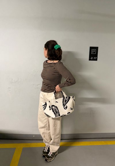 Joven con top marrón a rayas, pantalones blancos, zapatillas y un coletero verde sostiene una bolsa con patrón en blanco y negro en un aparcamiento.