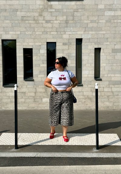 Camiseta blanca corta con gráfico de cereza roja, combinada con culottes de estampado de leopardo. Zapatos rojos, gafas de sol, de pie sobre un pavimento texturizado.