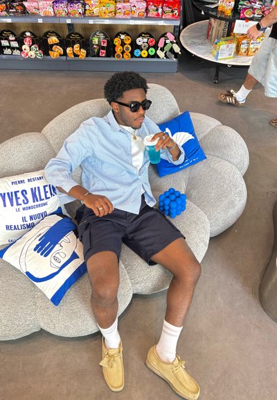 Sofá gris, redondeado, con tapicería texturizada; un cojín azul; una persona con gafas de sol y una camisa de cuadros azul claro bebe una bebida azul.