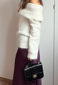 Maglione bianco a maglia con un design scollato sulle spalle, abbinato a una gonna viola scuro e una borsa nera in finta pelle di coccodrillo con dettagli dorati.