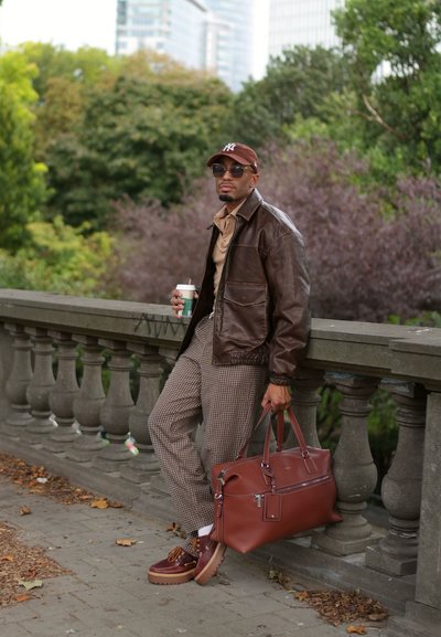 Chaqueta de cuero marrón sobre una camisa beige, pantalones de cuadros y bolsa marrón. Notables zapatos marrones con detalles y una taza de café en la mano.