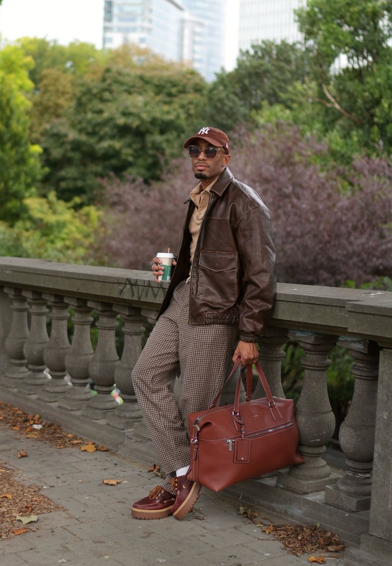 Giacca di pelle marrone sopra una camicia beige, pantaloni a scacchi e borsa marrone. Scarpe marroni notevoli con dettagli e una tazza di caffè in mano.