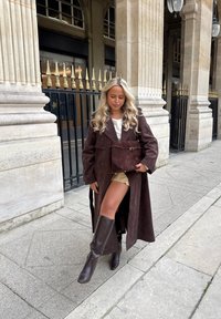 Lang brun semsket frakk med store slag, belte i midjen, kombinert med en beige miniskjørt og brune knelange støvler. Holder en matchende brun veske.