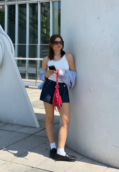 Mujer con gafas de sol sosteniendo un smartphone, apoyada en una pared blanca al aire libre, lleva una camiseta sin mangas blanca, falda azul marino y lleva un bolso pequeño rojo.