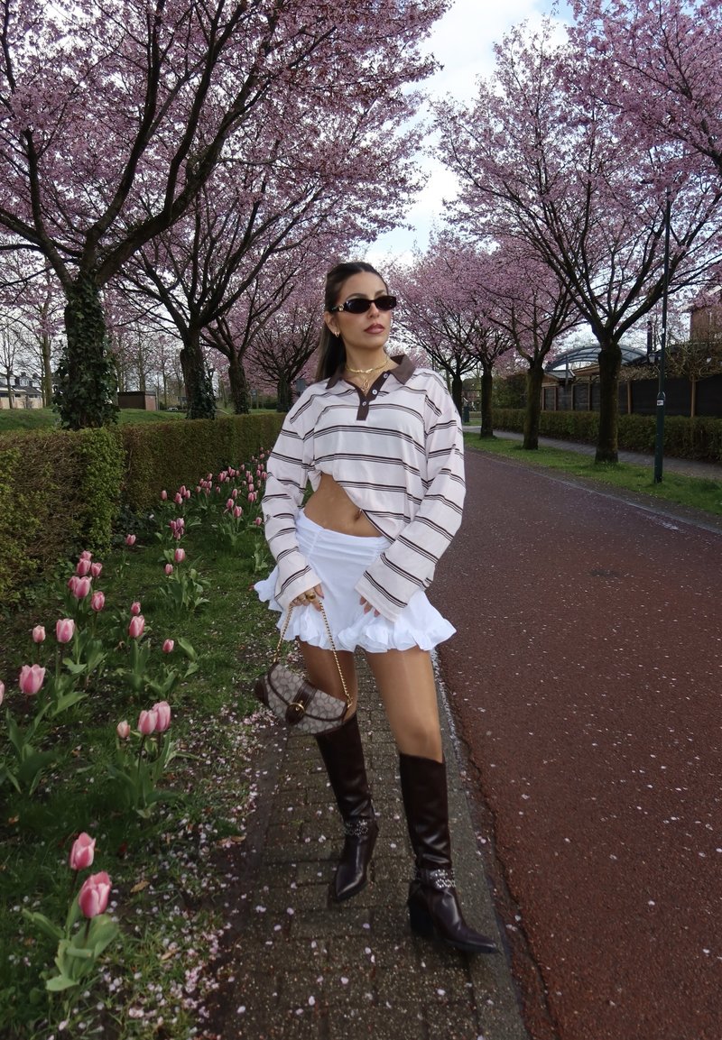 Femme portant des lunettes de soleil, une chemise à manches longues rayée, une jupe blanche et des bottes noires, se tenant sur un chemin bordé de tulipes roses et d’arbres en fleurs de cerisier.