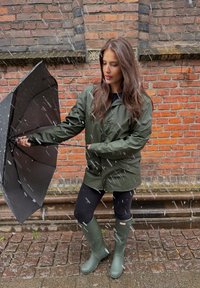 Cappotto impermeabile verde, ombrello nero con manico curvo, leggings neri e stivali di gomma verdi. Sfondo di muro di mattoni con neve.