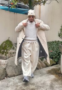 Manteau en laine crème, pull blanc en maille torsadée, pantalon beige clair et baskets grises. Coupe oversize, textures douces, accessoires métalliques, accents élégants.