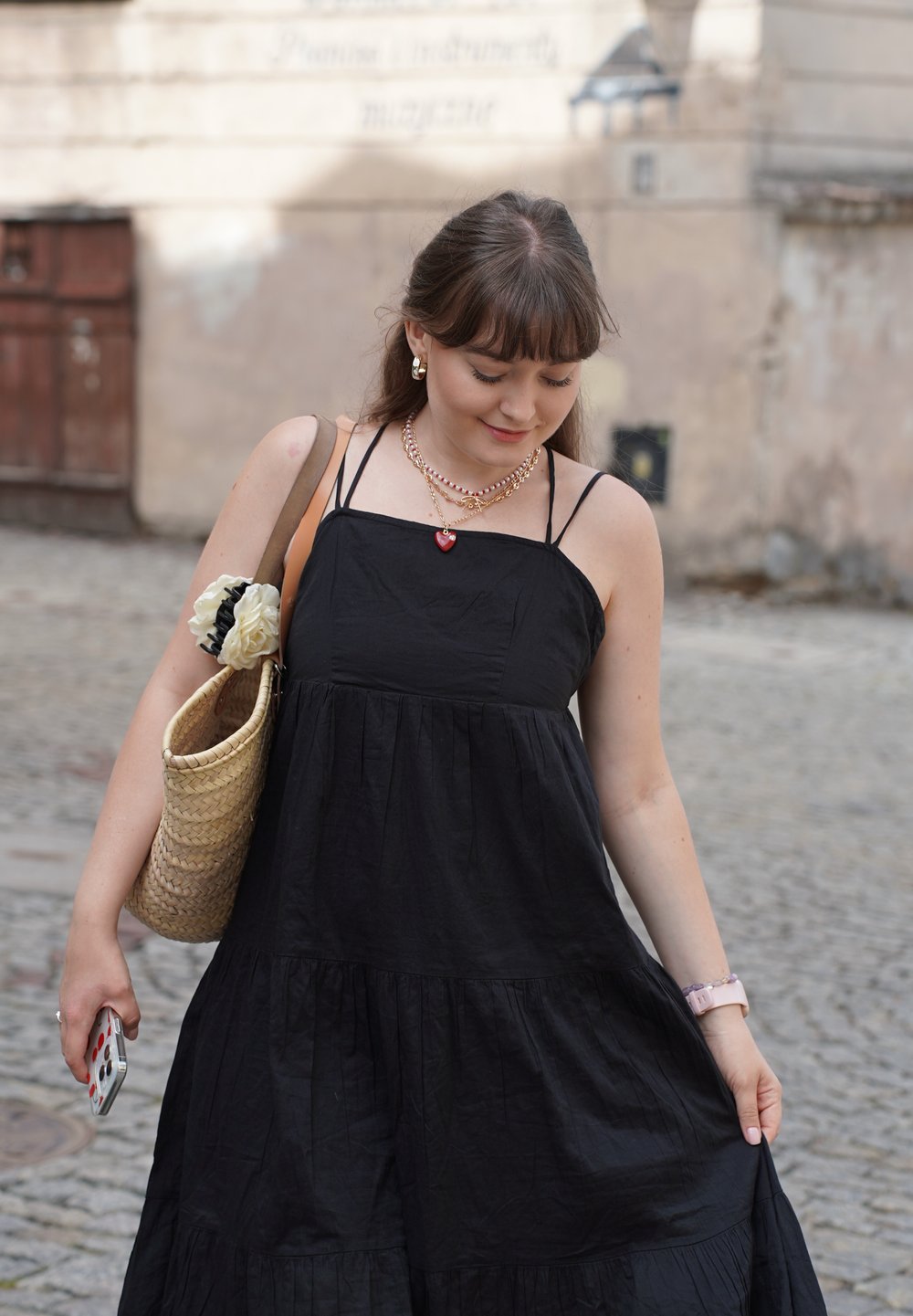 Abito neri senza maniche a strati in un materiale morbido, abbinato a una borsa di paglia intrecciata con un fiore decorativo. Telefono tenuto in mano.