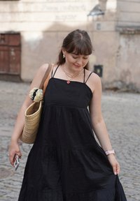 Abito neri senza maniche a strati in un materiale morbido, abbinato a una borsa di paglia intrecciata con un fiore decorativo. Telefono tenuto in mano.