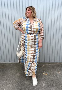 Maxi šaty s tie-dye vzorem v modré, hnědé a krémové barvě. Měkká tkanina, dlouhé rukávy a opasek v pase. Spárovány s bílými dřeváky a taškou.