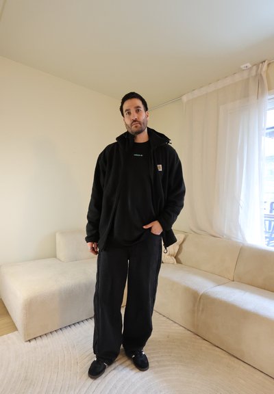 Hombre con chaqueta negra, camiseta negra de Adidas y pantalones negros sueltos, de pie en una sala de estar beige con sofá seccional y cortinas transparentes.