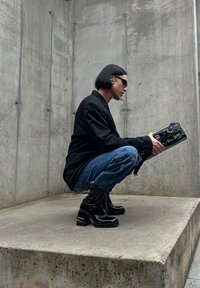 Casaco preto sobre uma blusa preta, jeans azuis e botas pretas de salto alto até o joelho. Segurando uma mala retangular preta com detalhes em metal.