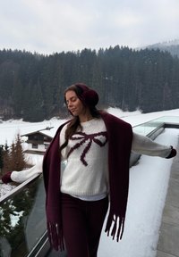 Jersey de punto con cuerpo blanco y diseño de lazo burdeos, combinado con una bufanda y orejeras a juego de color granate, en un entorno nevado al aire libre.