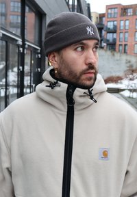 Άνδρας που φορά γκρι σκουφί και μπεζ μπουφάν με φερμουάρ με το λογότυπο Carhartt, κοιτώντας προς το πλάι σε αστικό υπαίθριο χώρο με κτίρια και χιόνι.