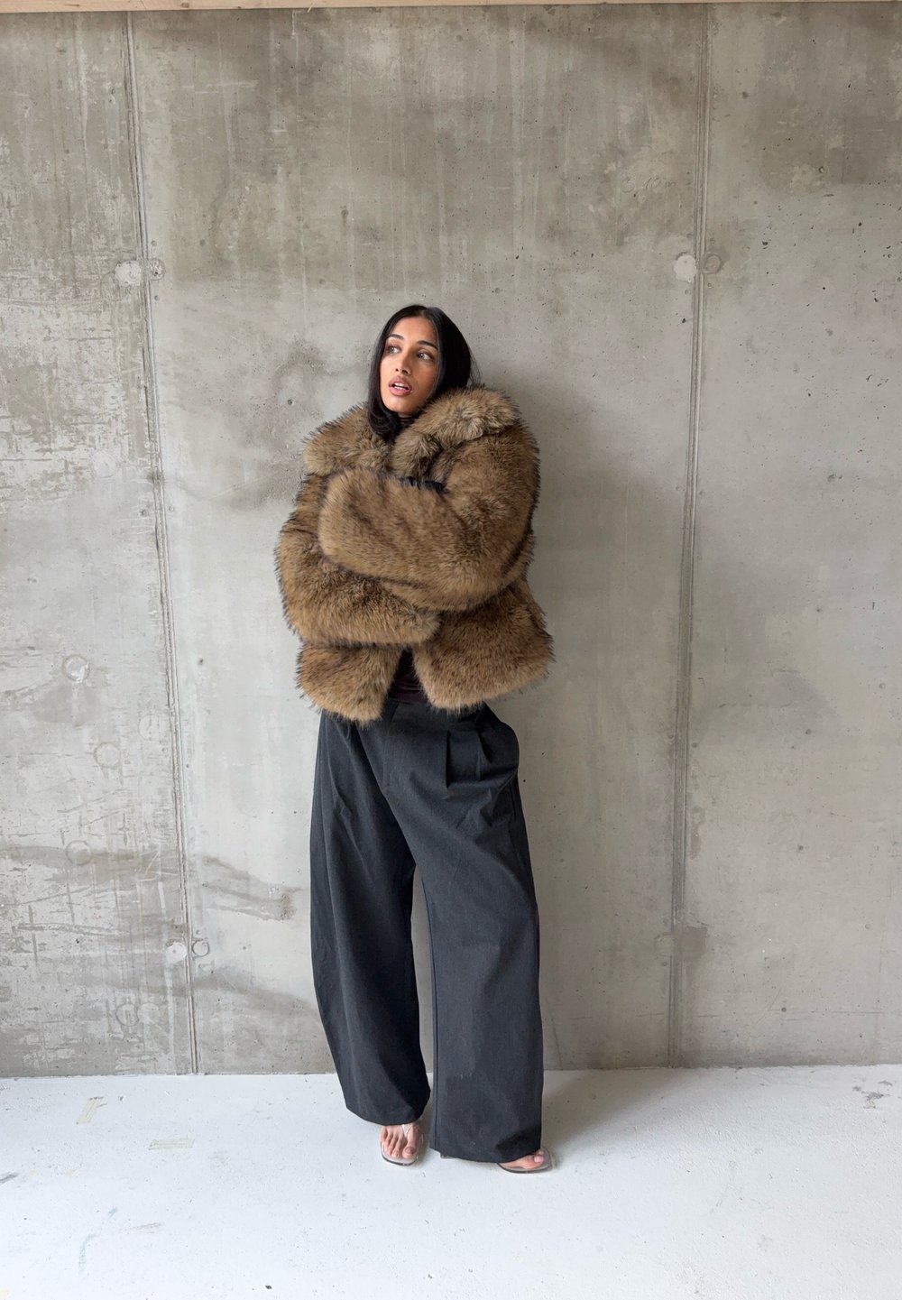Barna szőrmekabát széles gallérral, sötét felső fölött viselve, laza fekete nadrággal párosítva. Betonfal háttér. Egyszerű lábbeli.