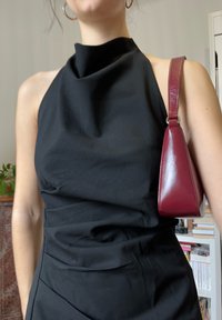 Femme portant une robe noire sans manches à col haut et un petit sac à bandoulière bordeaux sur son épaule droite.