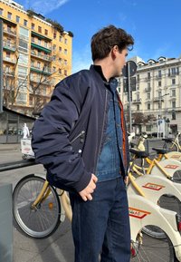 Ναυτικό bomber μπουφάν με ριγωτό γιακά και μανσέτες, μπροστινό φερμουάρ και τσέπες στα πλάγια, φορεμένο πάνω από ένα τζιν μπουφάν και σκούρα τζιν.