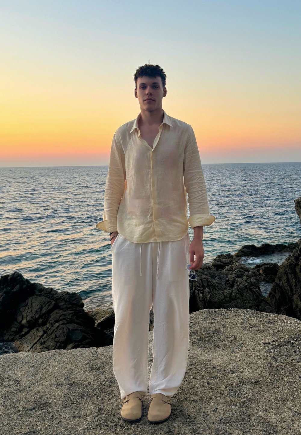 Ανοιχτό κίτρινο πουκάμισο από λινάρι με μακριά μανίκια, συνδυασμένο με λευκά παντελόνια με κορδόνι στη μέση και μπεζ παπούτσια, που στέκεται σε βραχώδη παραλία κατά τη διάρκεια της δύσης του ηλίου.