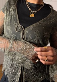 Olivi vihreä pitsillä päällystetty neuletakki, jossa on kukkakuviot, läpinäkyvät hihat, solmukiinnitys ja helmikoristelua. Päällä musta toppi.