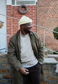 Palton verde peste un pulover bej, cu o căciulă tricotată galben deschis. Stă pe un zid de piatră lângă un zid de cărămidă și balustrade metalice.