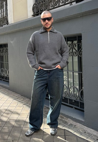 Suéter gris de canalé con cuello con cremallera, jeans holgados azules y zapatillas plateadas. Los accesorios incluyen gafas de sol oscuras.