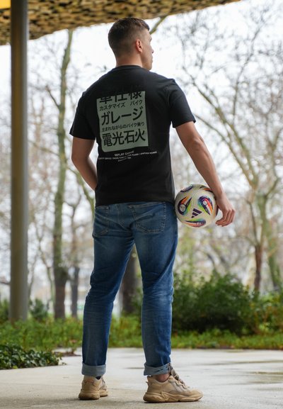 Camiseta negra con texto impreso en blanco y verde en la parte posterior, combinada con vaqueros azules y zapatillas beige. Sosteniendo un balón de fútbol multicolor.