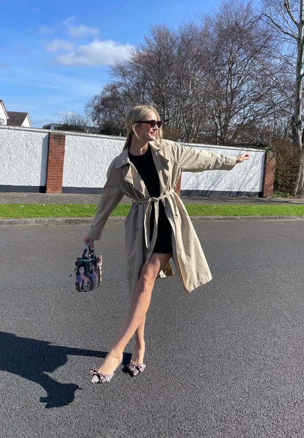 Manto bege com a cintura atada, vestido preto e sapatos com estampa de leopardo cor-de-rosa. A segurar uma mala de padrão colorido, em pé sobre asfalto.