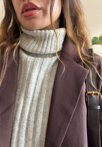 Brun blazer over en lys grå ribbet turtleneck sweater. Blazeren har strukturerede skuldre og et læderrem med en guldbuckle.