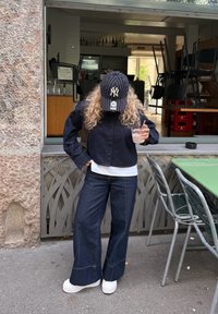 Person med lockigt hår som bär en randig basebollkeps, mörk jacka, vida jeans och vita sneakers, håller en drink utanför ett café.