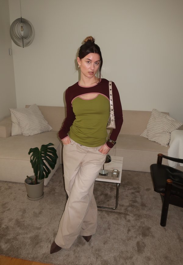 Frau mit Dutt-Frisur, die ein grünes und bordeauxfarbenes langärmeliges Oberteil und beige Hosen trägt, steht mit den Händen in den Taschen in einem Wohnzimmer.