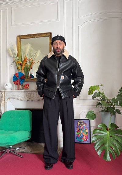 Hombre con chaqueta de cuero negra con cuello de borrego y boina negra, de pie en una habitación con una silla verde, plantas y obras de arte coloridas enmarcadas.