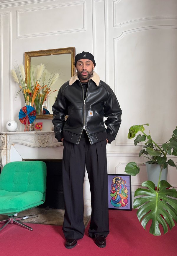 Mann trägt schwarze Lederjacke mit Lammfellkragen und schwarzen Barett, steht in einem Raum mit grünem Sessel, Pflanzen und bunten gerahmten Kunstwerken.