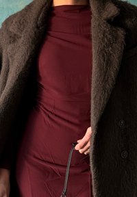 Personne portant un manteau duveteux marron foncé par-dessus une robe ajustée bordeaux, tenant une lanière en cuir noire dans la main droite.