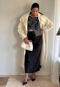 Hosszú krémszínű kabát, szürke argyle mintás kardigán, fekete szatén szoknya és fekete pántos magassarkú. Egyik kezében kis fehér kézitáskát tart.