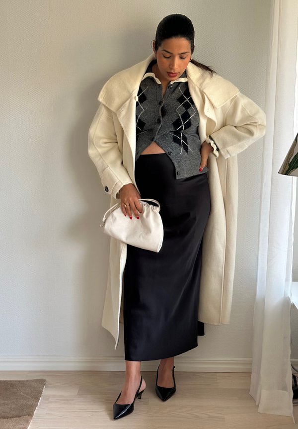 Lång krämfärgad kappa, grå kofta med argyle-mönster, svart sidenkjol och svarta slingback-klackar. En liten vit handväska hålls i ena handen.