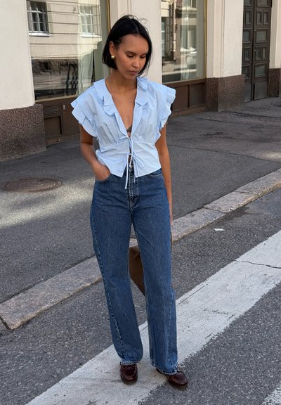 Blusa azul claro con volantes y cierre tipo lazo; combinada con jeans de mezclilla de pierna recta y cintura alta, y zapatos de piel marrón.