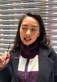 Viininpunaiset aurinkolasit pidettyinä suussa, yhdistettynä violetti-valkoiseen vetoketjulliseen neulepaitaan ja mustaan takkiin. Pitkät, stailatut kynnet korostavat ilmettä.