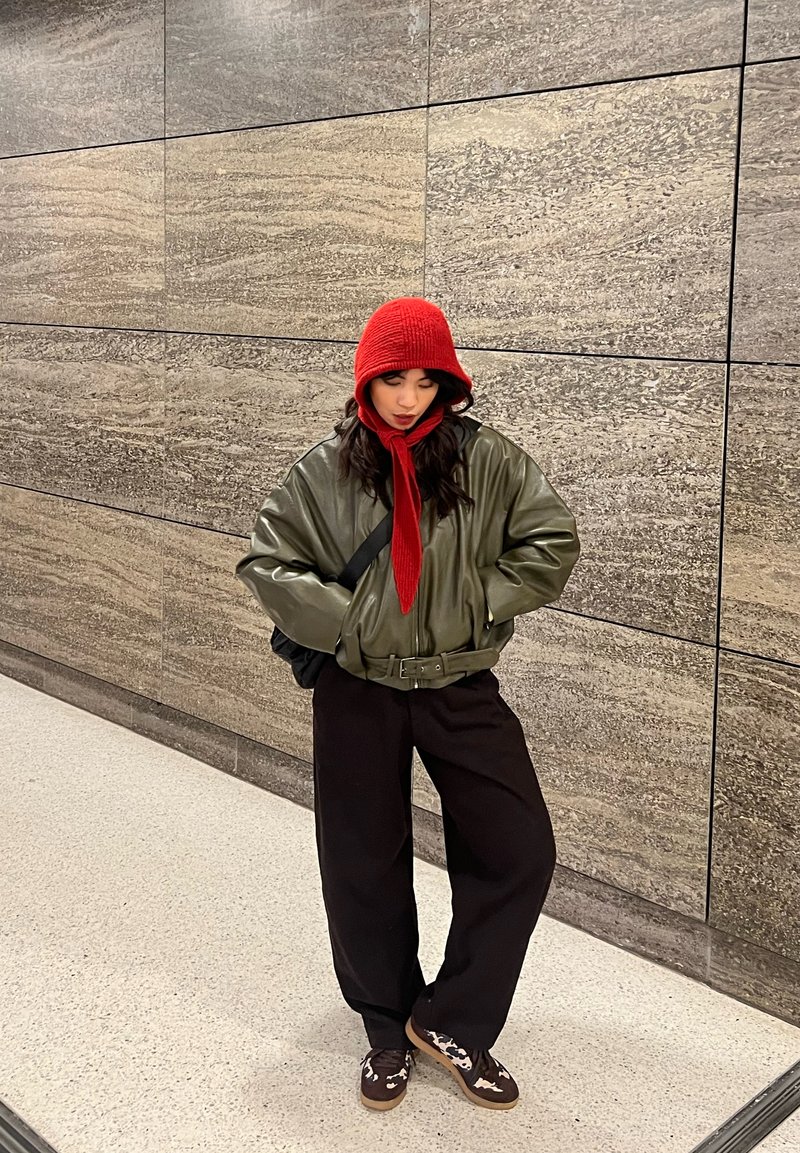 Grüne Leder-Bomberjacke mit Taillengürtel, roter Strickmütze und Schal, schwarze Hose mit weitem Bein, braune Sneakers mit weißen Akzenten.