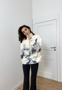 Fleece-lynlåsjakke i creme med blå abstrakte mønstre. Har høj krave, to frontlynlåse og tekstureret stof. Sorte bukser båret nedenunder.