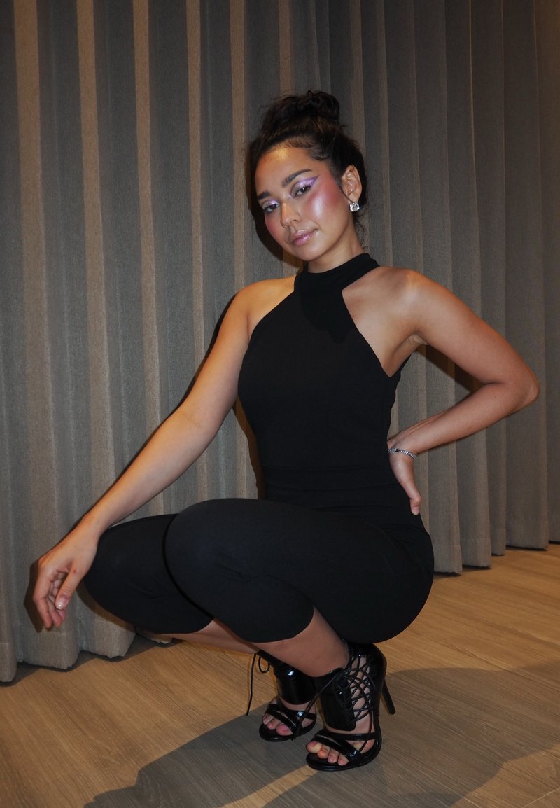 Jeune femme vêtue d'une tenue noire sans manches et de chaussures à talons hauts à lacets, accroupie à l'intérieur devant un rideau.