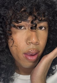 Lähiotoksen kuva kasvosta, jolla on kihara musta hius. Iho on vaaleanruskea, huulissa kiiltoa ja kultainen nenärenka. Kädentynkä lepää poskella.