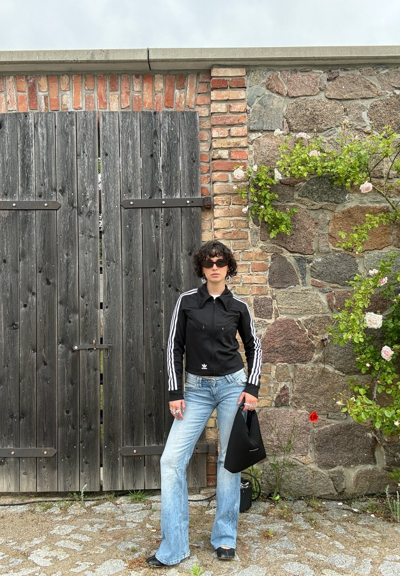 Personne portant des lunettes de soleil, une veste noire et un jean bleu se tient sur un chemin pavé près d'une porte en bois et d'un mur en pierre avec un buisson en fleurs.