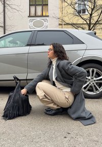 Femme aux cheveux bouclés et portant des lunettes accroupie à côté d'une voiture argentée, en train de fouiller dans un sac noir à franges, vêtue d'un manteau gris et d'un pantalon beige.