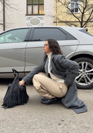 Mujer con cabello rizado y gafas agachada junto a un coche plateado, alcanzando una bolsa negra con flecos, vistiendo un abrigo gris y pantalones beige.