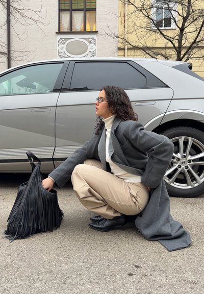 Mujer con cabello rizado y gafas agachada junto a un coche plateado, alcanzando una bolsa negra con flecos, vistiendo un abrigo gris y pantalones beige.