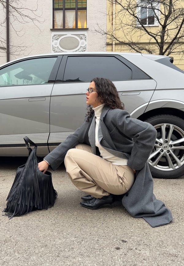 Femme aux cheveux bouclés et portant des lunettes accroupie à côté d'une voiture argentée, en train de fouiller dans un sac noir à franges, vêtue d'un manteau gris et d'un pantalon beige.